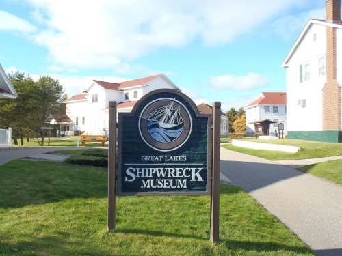 Sign for the Great Lakes Shipwreck Museum, surrounded by green grass and buildings under a blue sky.