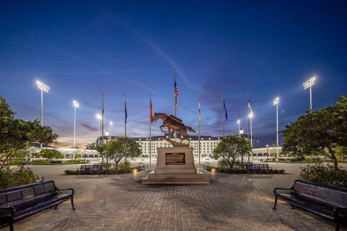 A bronze horse statue at dusk, surrounded by flags and illuminated by stadium lights, with benches in the foreground.