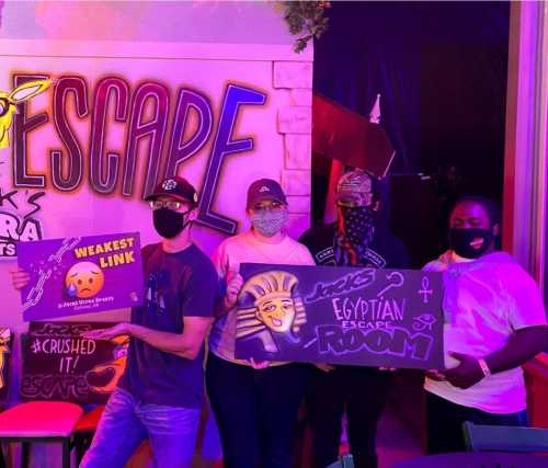 Four people in masks hold signs at an escape room event, with colorful decorations in the background.