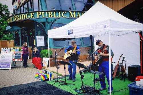 Two musicians perform under a tent outside a public market, while a few people enjoy the scene nearby.