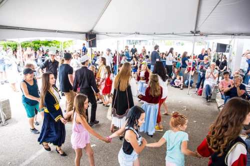 A lively crowd gathers under a tent as people dance in traditional costumes, enjoying a festive event.