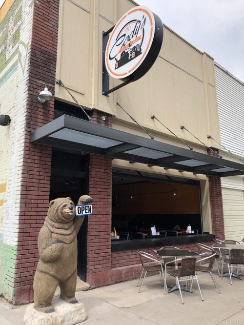 A restaurant exterior with a bear statue holding an "OPEN" sign and outdoor seating.