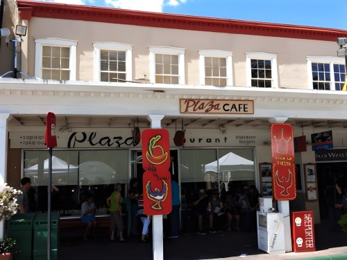 Exterior of Plaza Cafe with outdoor seating, colorful decorations, and a bright blue sky above.