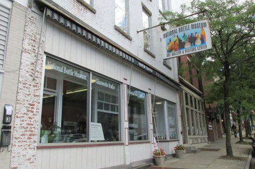 Exterior of the National Bottle Museum, featuring a sign and large windows, with trees lining the sidewalk.