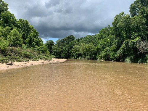 A calm river flows through a lush green landscape under a cloudy sky.
