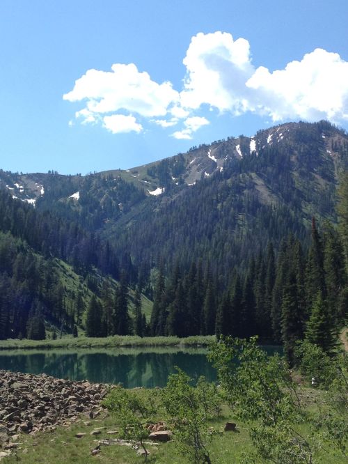 A serene mountain landscape with a clear lake, surrounded by lush green trees and snow-capped peaks under a blue sky.