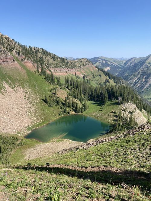 A serene mountain landscape featuring a clear green lake surrounded by lush greenery and rugged hills under a blue sky.