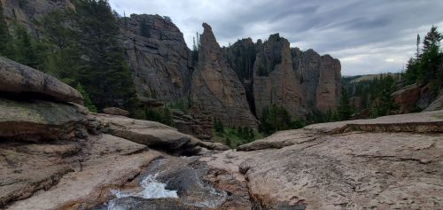 A rocky landscape with towering cliffs and a small stream flowing through, surrounded by trees under a cloudy sky.