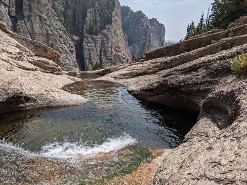 A serene rocky landscape with a clear pool of water surrounded by cliffs and trees under a hazy sky.