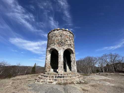 Stone tower with arched openings, set against a blue sky and sparse trees on a rocky landscape.