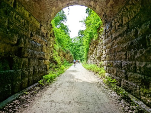 A stone archway frames a dirt path surrounded by lush greenery, with people walking in the distance.