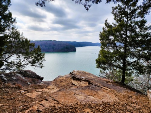 A serene view of a lake surrounded by trees and rocky terrain under a cloudy sky.