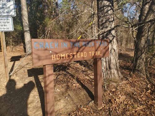 Signpost for "Crack in the Rock Trail" and "Homestead Trail" in a wooded area with trees and fallen leaves.