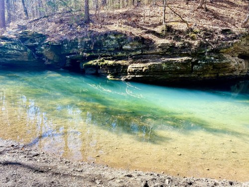 A serene river with clear turquoise water flows beside rocky banks, surrounded by trees and sunlight.