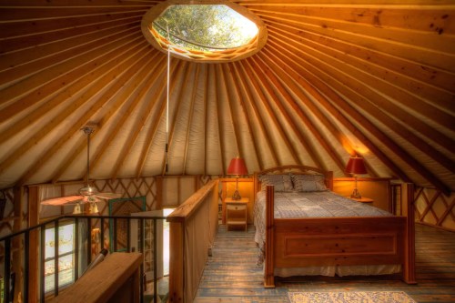 Cozy wooden yurt interior with a circular skylight, a bed, and warm lighting.