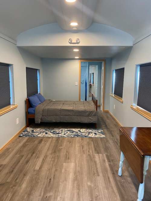 A cozy bedroom with a bed, blue bedding, wooden furniture, and two windows with blinds, featuring a light wood floor.