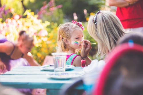 A young girl with a bow in her hair gets her face painted while sitting at a table in a colorful garden.