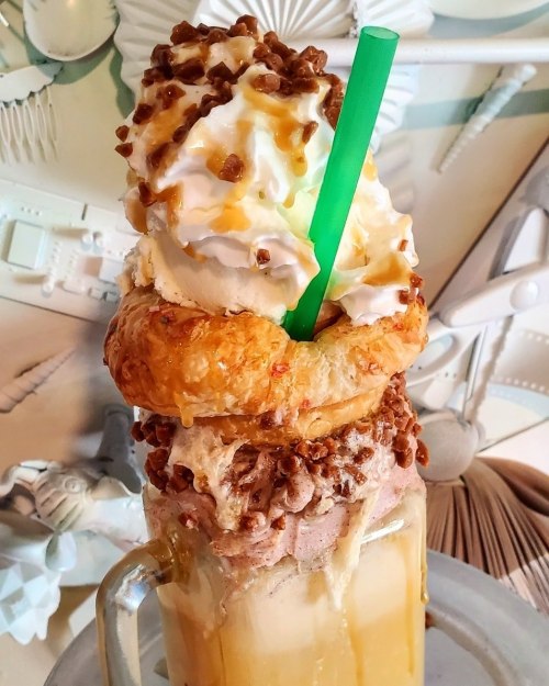 A towering dessert drink with whipped cream, caramel, and a croissant on top, served in a glass with a green straw.