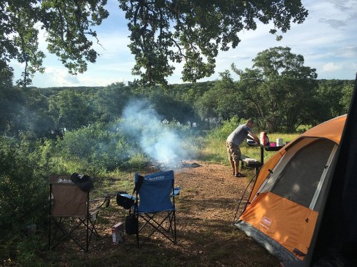 A campsite with two chairs, a tent, and a person cooking over a fire, surrounded by trees and a scenic view.