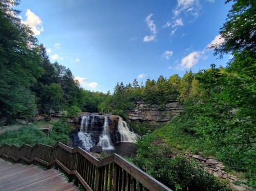 A scenic view of a waterfall surrounded by lush greenery and a wooden walkway under a blue sky with scattered clouds.