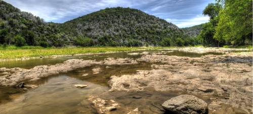 A serene river scene with rocky banks, lush greenery, and rolling hills under a clear blue sky.