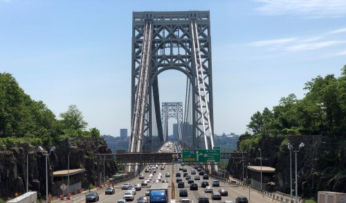The George Washington Bridge spans a river, with heavy traffic and greenery on either side under a clear blue sky.