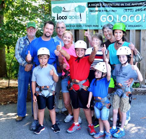 A group of adults and children in helmets giving thumbs up, standing in front of a sign for Loco Ropes in Arkansas.
