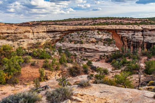 A natural stone arch spans a canyon, surrounded by rugged terrain and greenery under a partly cloudy sky.