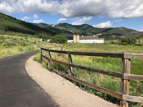 A winding path leads through green fields, with mountains in the background and a white building near a wooden fence.