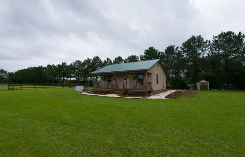A small house with a green metal roof surrounded by a grassy yard and trees under a cloudy sky.