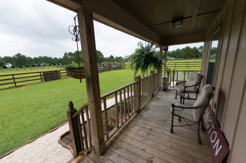 A wooden porch with two chairs overlooks a grassy yard and fenced area under a cloudy sky.