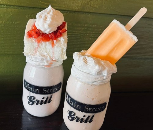 Two milkshakes in jars topped with whipped cream, one with a slice of cake and the other with a popsicle.