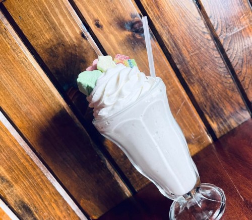 A tall glass of milkshake topped with whipped cream and colorful marshmallows, set against a wooden background.
