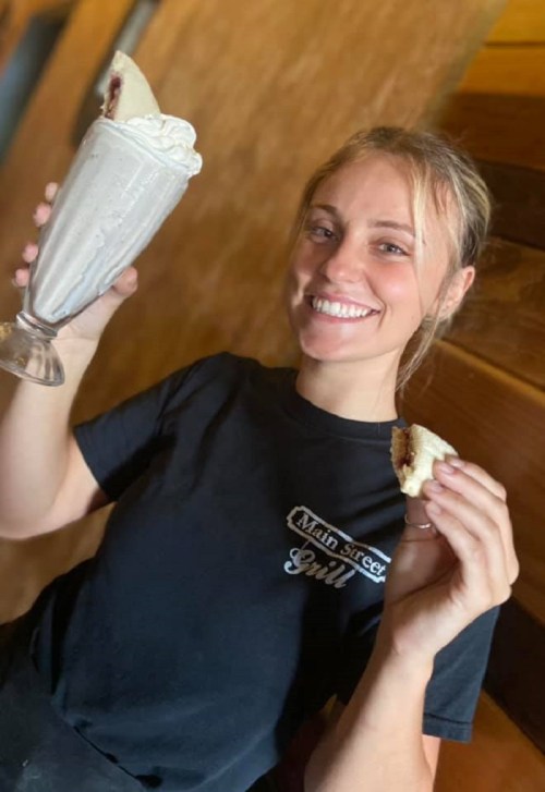 A smiling young woman holds a milkshake in one hand and a dessert in the other, wearing a black shirt.