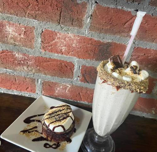 A decadent milkshake topped with marshmallows and a chocolate dessert on a plate, set against a brick wall.