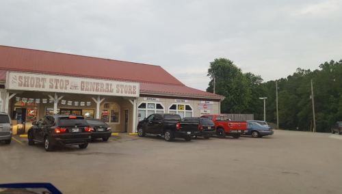 A general store with a red roof, surrounded by parked cars, set against a cloudy sky and green trees.
