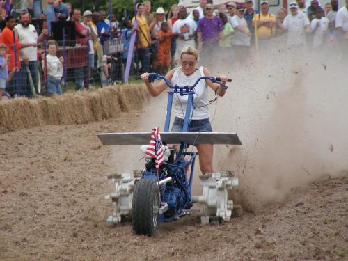 A woman operates a plow in a dirt field, surrounded by a cheering crowd, with dust flying around her.