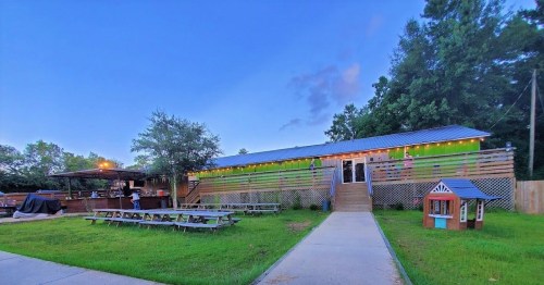 A colorful outdoor restaurant with picnic tables, a bar area, and a grassy lawn under a blue sky at dusk.