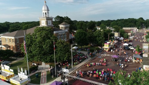 Aerial view of a lively town square with a clock tower, surrounded by vendors and people enjoying a festival.
