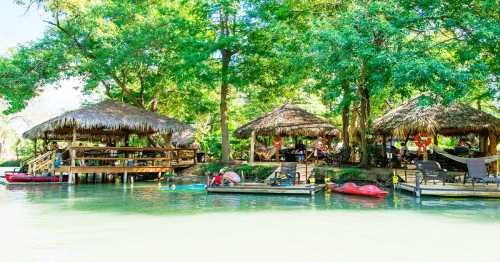 Lush green landscape with thatched-roof huts by a calm river, featuring kayaks and people enjoying the serene environment.