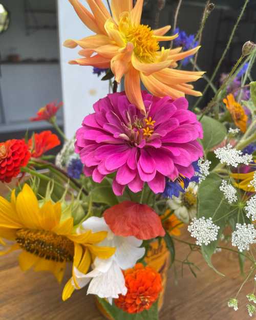 A vibrant bouquet of mixed flowers, featuring pink zinnias, orange dahlias, sunflowers, and delicate white blooms.