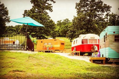 Colorful vintage trailers lined up on a grassy area, with a blue umbrella providing shade. Trees in the background.