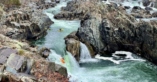 Two kayakers navigate rocky rapids with a small waterfall in a scenic river landscape.