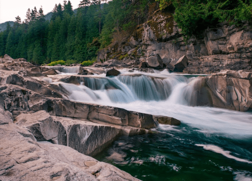 A serene river flows over smooth rocks, surrounded by lush green trees and a rocky landscape.