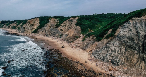 Aerial view of a rocky beach with cliffs and green hills, waves gently lapping at the shore.
