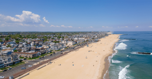 Aerial view of a beachside town with sandy shores, ocean waves, and residential buildings under a clear blue sky.