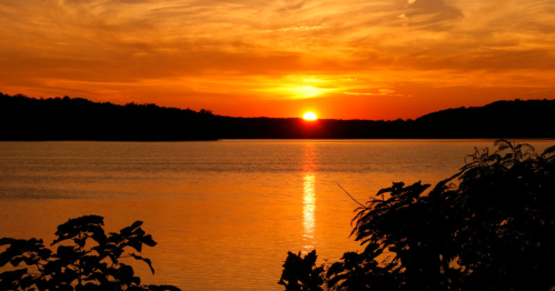 A vibrant sunset over a calm lake, with silhouettes of trees in the foreground and colorful clouds in the sky.