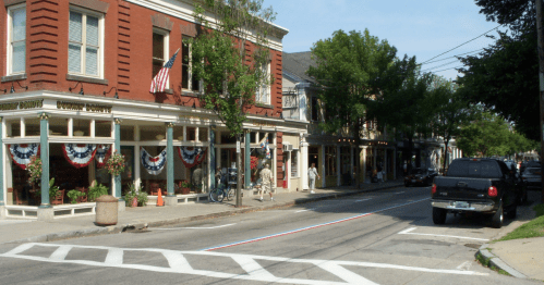 A charming street scene featuring brick buildings, American flags, and festive bunting, with parked cars and trees lining the road.