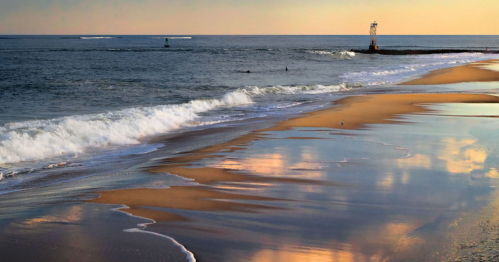 A serene beach at sunset, with gentle waves, a lighthouse in the distance, and colorful reflections on the wet sand.