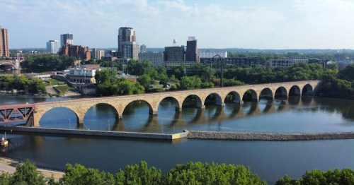 Aerial view of a stone arch bridge spanning a river, with city buildings and greenery in the background.